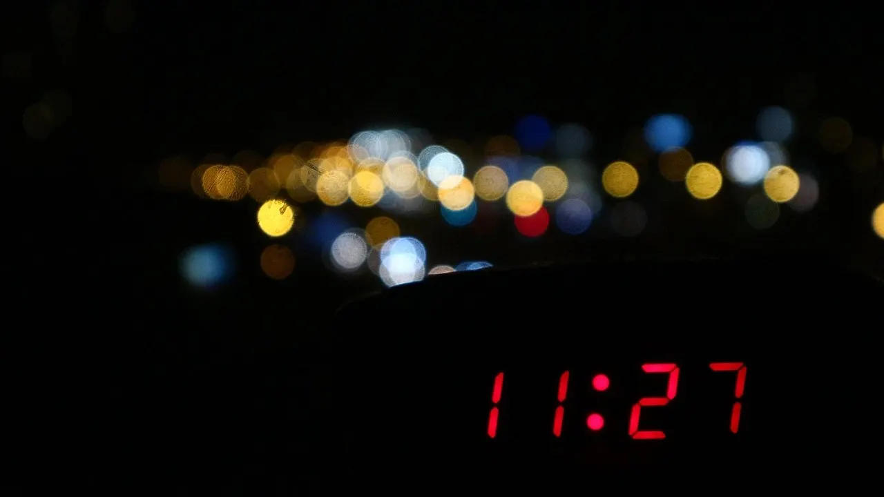 A red digital clock glowing in the dark against blurred city lights at night.