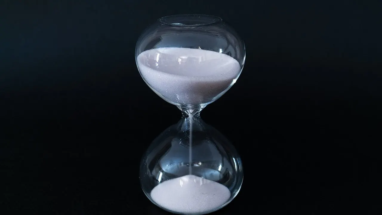 A glass hourglass on a black background with sand trickling through: a schedule counting down to a run that may never fire.